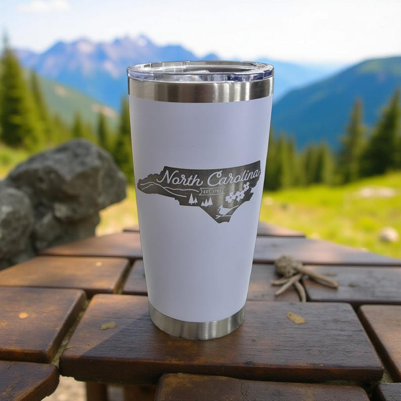 May include: A white stainless steel tumbler with a silver rim and lid. The tumbler features a gray outline of North Carolina with the text "North Carolina" and other graphics. The tumbler is sitting on a wooden table.