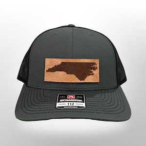 May include: A gray and black trucker hat with a brown leather patch featuring the outline of North Carolina. The hat has the text "Richardson" and "Original 112 Trucker" on the front.