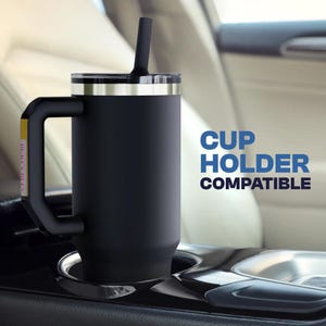 May include: A black insulated travel mug with a handle and a straw. The mug has a silver rim and a black lid. The text "CUP HOLDER COMPATIBLE" is in blue. The mug is in a car cup holder.
