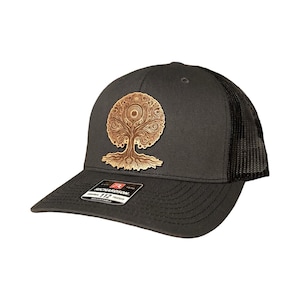 May include: A black and black mesh trucker hat with a gold-colored tree of life design. The hat has a black adjustable strap with a "112" label.
