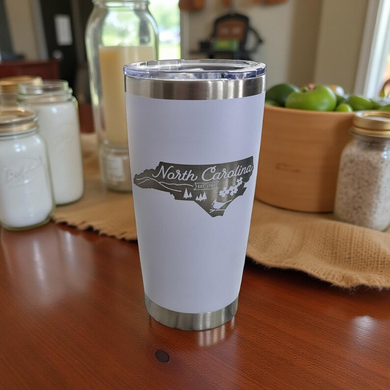 May include: A white stainless steel tumbler with a silver rim and base. The tumbler features a gray outline of North Carolina with the words "North Carolina" and other text within the state's shape.