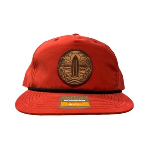 May include: Red baseball cap with a black brim and a brown leather patch featuring a surfboard and palm trees. The patch has the text "RICHARDSON" on it.