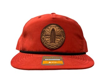 May include: Red baseball cap with a black brim and a brown leather patch featuring a surfboard and palm trees. The patch has the text "RICHARDSON" on it.