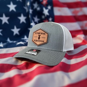 May include: A gray trucker hat with a white mesh back and a curved brim. The hat features a brown patch with the text "12 DAY WAR SURVIVOR" and a bomb graphic. The hat is set against an American flag.