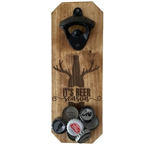 May include: Wooden bottle opener with a black metal bottle opener and a deer silhouette with the text "It's Beer Season". The bottle opener has a magnetic back to hold bottle caps. There are several bottle caps attached to the back of the bottle opener.