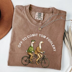 May include: A brown Comfort Colors t-shirt with the phrase "OFF TO COMIT TOM FOOLERY" in a semi-circle. The shirt features a tandem bicycle with two geese wearing clothing. A silver necklace is draped across the shirt.