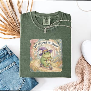 Può includere: T-shirt verde oliva con una grafica stravagante di una rana con un cappello da strega su uno scooter, con la scritta "Off to Commit Tom Foolery". Sono visibili anche una collana d'argento e scarpe bianche.