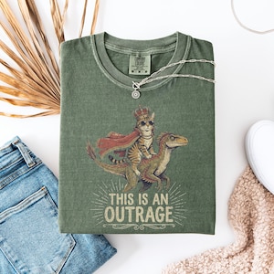 Peut inclure: T-shirt vert avec un motif d'un chat couronné et capé chevauchant un dinosaure, avec le texte "THIS IS AN OUTRAGE". Le t-shirt est plié avec un collier argenté, un jean bleu et des baskets blanches.