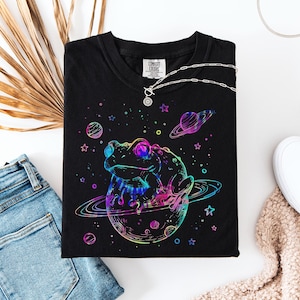 Peut inclure: Un t-shirt noir avec un motif graphique aux couleurs néon représentant une grenouille assise sur une planète entourée d'étoiles et d'autres corps célestes. Le design utilise des contours bleu, rose et violet. Un collier en argent est posé sur le col.