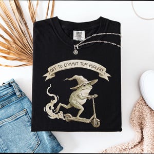 Puede incluir: Camiseta negra con un diseño de una rana con sombrero de mago en un patinete, con el texto "OFF TO COMMIT TOM FOOLERY". Un collar plateado y unos vaqueros azules están junto a la camiseta. Ropa informal.