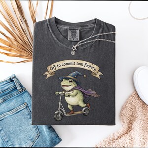 Può includere: T-shirt grigio scuro con un'illustrazione di una rana su uno scooter, con un cappello da mago e un mantello. Lo striscione recita "Off to commit tomfoolery". Sono visibili anche una collana d'argento e scarpe bianche.