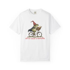 May include: White t-shirt featuring a whimsical illustration of a frog riding a bicycle while wearing a wizard hat and cape, holding a book. The text "off to cause a kerfuffle" is printed below the image.