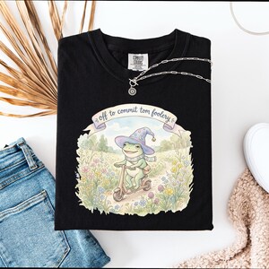 Può includere: T-shirt nero con un'illustrazione di una rana con cappello da mago su uno scooter, con la scritta "off to commit tom foolery". Sono visibili anche una collana d'argento e jeans blu.