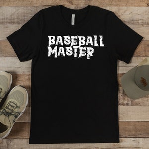 Op de afbeelding: Zwart T-shirt met de tekst "BASEBALL MASTER" in versleten witte letters. De afbeelding toont ook een paar beige sportschoenen met grijze veters en een grijze baseballpet met een bruin embleem.
