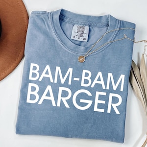 May include: A blue t-shirt with the text "BAM-BAM BARGER" in white block letters. A gold necklace is draped across the folded shirt. A brown hat and dried plant are in the background.