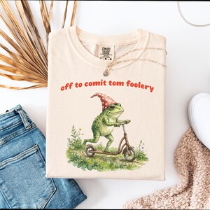 Può includere: T-shirt color crema con la scritta rossa "off to comit tom foolery". L'immagine mostra una rana verde con un cappello a pois rossi e bianchi, su uno scooter.