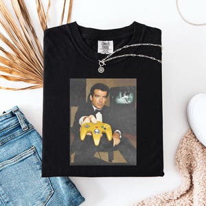 May include: Black t-shirt featuring a graphic of a man in a tuxedo holding a yellow video game controller. The image on the shirt shows a retro video game scene. The shirt has the text "COMFORT COLORS" on the tag.