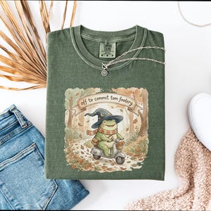 Può includere: T-shirt verde con un'illustrazione di una rana su uno scooter, con un cappello da mago e una sciarpa. Il testo sulla maglietta recita "off to commit tom foolery". Sono visibili anche una collana d'argento e scarpe bianche.