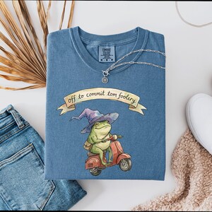 Può includere: Maglietta blu con una grafica di una rana su uno scooter rosso con un cappello da mago. Il testo sulla maglietta recita "off to commit tom foolery". Una collana d'argento e scarpe bianche sono anche nell'immagine.