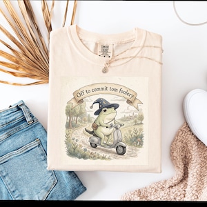 May include: A cream-coloured t-shirt with a cartoon frog on a scooter, wearing a wizard hat, and a banner that reads "Off to commit tom foolery." The shirt is styled with blue jeans and white trainers.