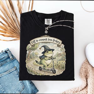 Peut inclure: T-shirt noir avec une illustration d'une grenouille en chapeau de sorcier et cape, sur un scooter. Le texte "Off to commit tomfoolery" est sur une bannière. Un collier argenté et des chaussures blanches sont visibles.