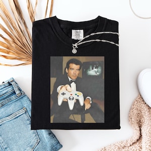 Peut inclure: T-shirt noir avec une image rétro d'un homme tenant une manette de jeu vidéo. L'image est imprimée sur un t-shirt noir. Le t-shirt a un col rond et des manches courtes. Le texte "COMFORT COLORS" est imprimé au dos du t-shirt.