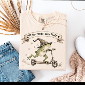 May include: Cream-colored t-shirt featuring a whimsical illustration of a green frog wearing a wizard hat and riding a scooter. The shirt has the text "Off to commit tom foolery" on a banner. The shirt is folded and placed with blue jeans and white shoes.