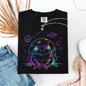 Peut inclure: Un t-shirt noir avec un motif graphique coloré de style néon représentant une grenouille assise devant une grande lune, entourée de petites planètes et d'étoiles. Le design utilise des contours vifs roses, bleus et verts sur le tissu.