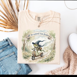 Peut inclure: T-shirt crème avec une illustration à l'aquarelle fantaisiste d'une grenouille sur un scooter dans une forêt. La grenouille porte un chapeau de sorcier et le texte dit "off to commit tom foolery."