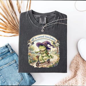 Può includere: T-shirt grigio scuro con una rana con cappello da mago su uno scooter, con la scritta "off to commit tomfoolery". La maglietta è stesa con jeans blu, scarpe bianche e una collana. La maglietta è di cotone.