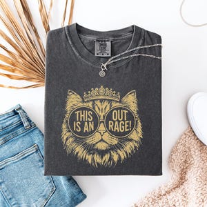 Peut inclure: T-shirt gris foncé avec un motif de chat doré portant des lunettes et une couronne. Le texte sur le t-shirt se lit "THIS IS AN OUT RAGE!". Le t-shirt est plié à côté d'un jean bleu, d'un collier argenté et de baskets blanches.
