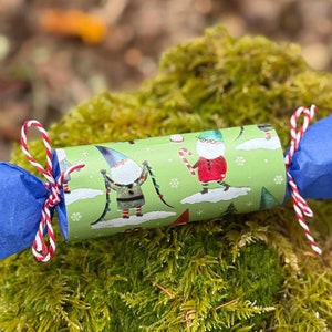 May include: A blue and white paper tube with a red and white string tied around it. The tube is decorated with a green background and a pattern of gnomes skiing.