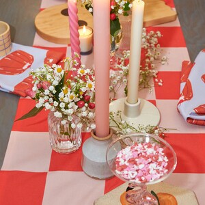 May include: A table setting with a red and pink checkered tablecloth. There are three candles, two pink and one white, in candle holders. There are also small white flowers in a glass vase and a glass bowl filled with pink and white sprinkles.