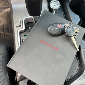 May include: A gray vehicle log book with the words "VEHICLE LOG" printed in red on the cover. A black car key with a red button is on top of the book, along with a silver key with a black Toyota logo.