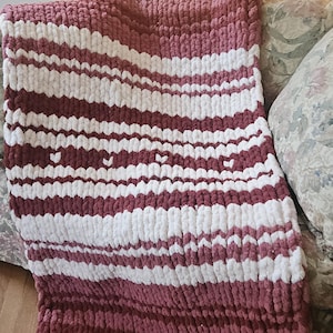 May include: A chunky knit throw blanket featuring alternating horizontal stripes of dusty rose and white yarn. The texture is created with thick, soft loops of yarn, and a subtle pattern of small heart shapes is integrated into the design.