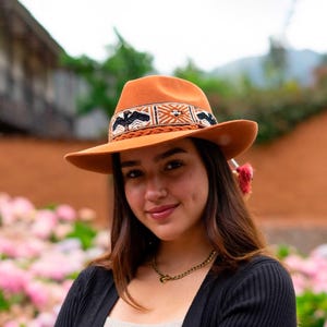 May include: A burnt orange fedora hat with a decorative band featuring black and white geometric patterns. The hat has a wide brim and is likely made of felt. The hat is part of the Handmade Alpaca Peru collection.