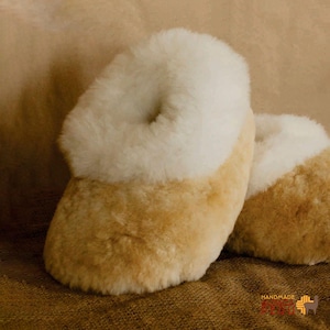 May include: A pair of cozy, handmade slippers. The slippers are a light tan color with a fluffy, off-white trim around the opening. The slippers appear soft and warm, perfect for indoor wear.