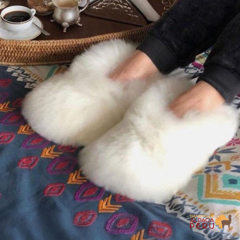 May include: Fluffy white alpaca wool slippers. These cozy slippers are designed for indoor use and are made from soft, natural alpaca wool. The slippers are shown on a patterned blue and teal fabric.