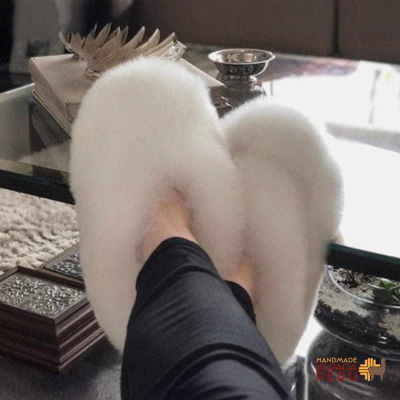 May include: Fluffy white slippers made of soft material. The slippers are a light, neutral color and appear to be comfortable. The slippers are a cozy footwear option for indoor use.