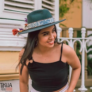 May include: A woman wearing a teal fedora with a blue and white patterned band and a black tank top. The hat has a tassel with red, orange, and purple accents. The woman is smiling and looking to the right.