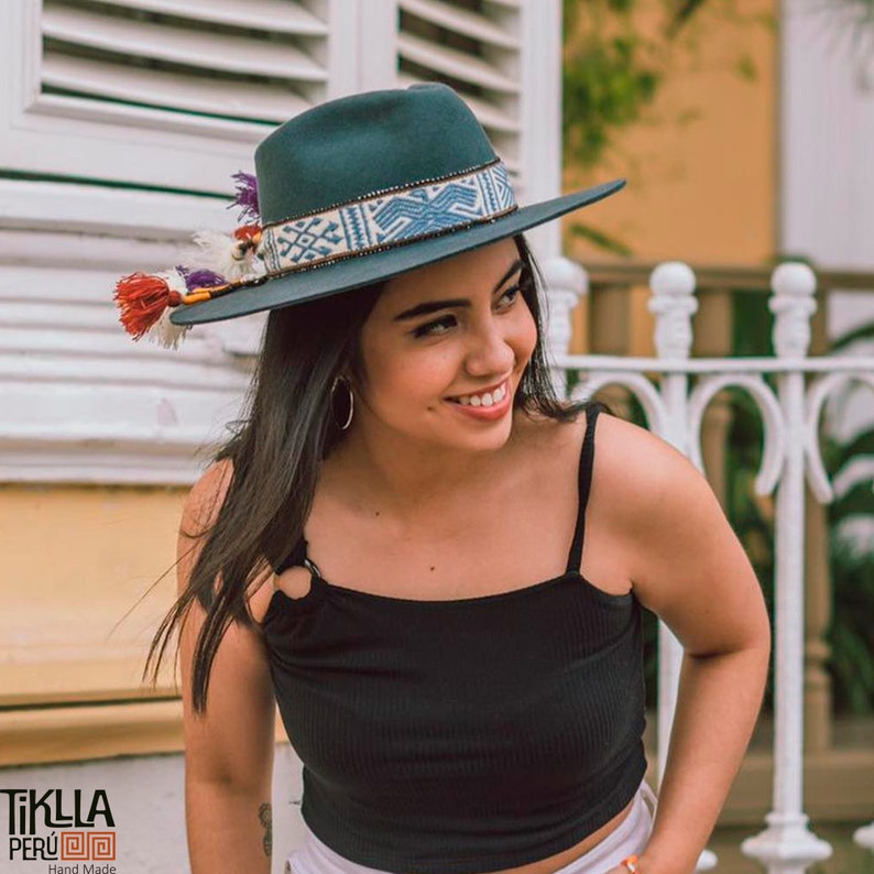 May include: A woman wearing a teal fedora with a blue and white patterned band and a black tank top. The hat has a tassel with red, orange, and purple accents. The woman is smiling and looking to the right.