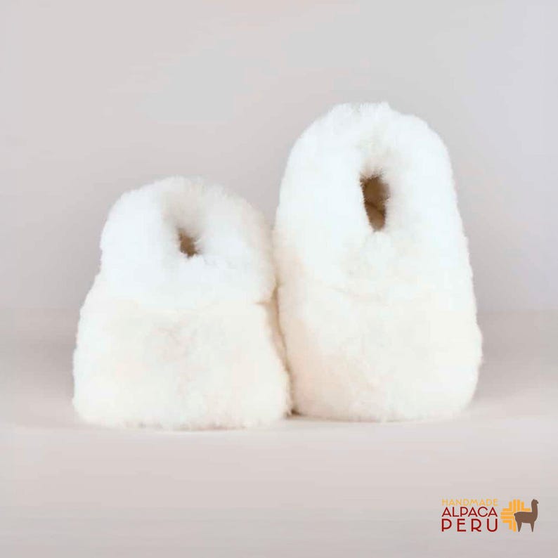 May include: Two fluffy, off-white alpaca wool slippers. The slippers have a rounded shape with a hole at the top for the foot. The slippers are soft and plush, perfect for indoor wear. The slippers are handmade.