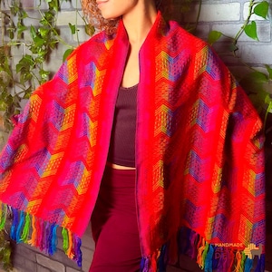 May include: A vibrant red shawl with a geometric pattern in shades of yellow, blue, and purple. The shawl has a fringed edge with colorful tassels. The handmade textile is draped over the shoulders.
