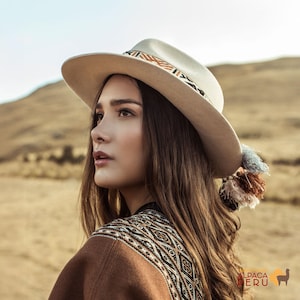 Può includere: Un cappello fedora beige con una fascia decorativa con motivi geometrici marroni, arancioni e neri. Il cappello ha un'ampia tesa e un nappa. È visibile anche uno scialle marrone fantasia.