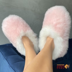 May include: Pink and white fluffy alpaca fur slippers. The slippers have a soft, plush appearance with a light pink upper and white trim. The slippers are handmade in Peru.