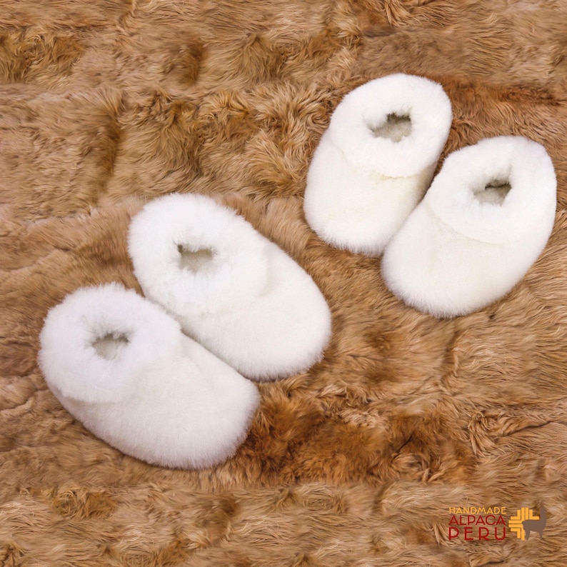 May include: Four pairs of white alpaca wool slippers are arranged on a brown, textured surface. The slippers have a soft, fluffy appearance, with a rolled cuff around the opening. The slippers are designed for indoor wear and are likely warm and comfortable.