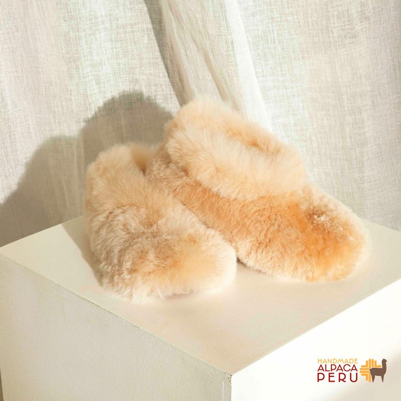 May include: A pair of light tan, fluffy slippers made of alpaca wool. The slippers are resting on a white surface, with a soft, neutral-toned background. The slippers have a plush, textured appearance, suggesting warmth and comfort. The image includes the text "HANDMADE ALPACA PERU".