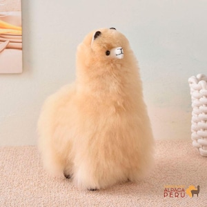 May include: A plush, light tan alpaca stuffed animal with a fluffy, textured coat. The alpaca has black ears, a white muzzle, and small black feet. The Alpaca Peru logo is visible in the lower right corner.