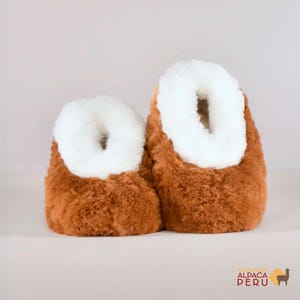 May include: A pair of brown and white alpaca wool slippers. The slippers have a fluffy, textured appearance with a brown exterior and a white interior lining. The slippers are designed to keep feet warm and comfortable.