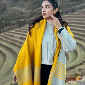 Puede incluir: Una mujer lleva un chal amarillo y gris estampado sobre una chaqueta gris. El chal tiene un diseño geométrico en gris. La imagen está ambientada sobre un fondo de colinas aterrazadas. El texto "HANDMADE ALPACA PERU" es visible.